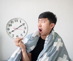 Asian man with blanket cover body wake up late feel shocked and look at clock in hand on white isolated
