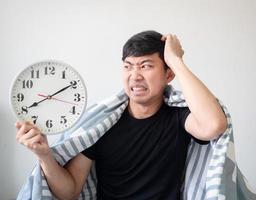 el hombre se despierta tarde, se siente sorprendido, toca la cabeza con la mano con el cuerpo cubierto de una manta y mira el reloj en la mano foto