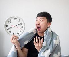 Asian man shocked face and looking at clock in hand and hug pillow on white isolated wake up late concept photo