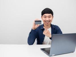 el hombre asiático se sienta con la computadora portátil en la mesa y señala con el dedo la tarjeta de crédito en la mano foto