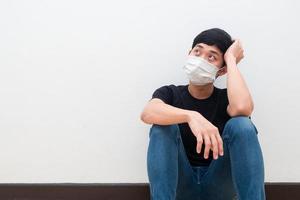 Drepressed Asian man wearing mask sit on the floor touch his head and looking up white wall background