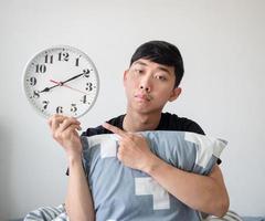 Young man with pillow point finger at clock in hand and feel bored on white isolated work late concept photo