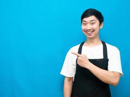 Asian man wearing apron happy smile point finger at copy space blue background photo