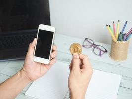 teléfono inteligente de mano y bitcoin dorado con espacio de trabajo en el fondo de la mesa de madera blanca, teléfono y bitcoin en la mano y gafas de computadora portátil en el fondo del escritorio foto