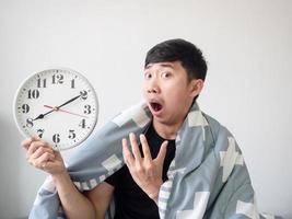 Asian man wake up with blanket cover body and feel bored at face look at clock in hand on white isolated photo