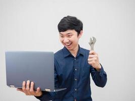 hombre asiático sosteniendo una llave ajustable y mirando la computadora portátil en su mano cara confiada para fijarse en el fondo blanco foto