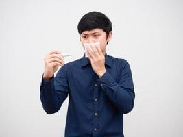 hombre asiático que se siente enfermo cierra la nariz con papel tisú y mira el termómetro en la mano cara seria fondo blanco foto