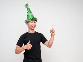 joven asiático cara feliz mira la cámara con sombrero verde apuntando con el dedo derecho sobre el pulgar hacia arriba y una sonrisa feliz sobre fondo blanco aislado foto