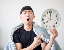 Man wake up with blanket cover body feel sleepy and yawn point finger at clock in hand on white isolated,Sleepy man concept photo