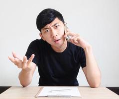 Young man black shirt sit at the desk talking with phone feel serious and confused face look at camera with clipboard on the desk,Business concept photo