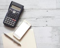 pluma plateada de pantalla blanca de teléfono inteligente en un cuaderno de página en blanco con calculadora en la vista superior de la mesa de madera blanca y espacio de copia foto