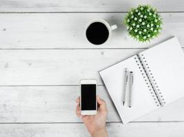 Workspace on white wood table top view copy space,Silver pen on blank page notebook and smartphome black screen in hand with coffee cup minimal concept photo