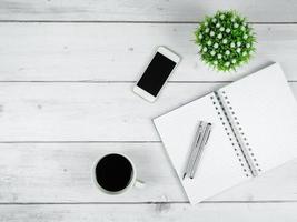 espacio de trabajo en el espacio de copia de la vista superior de la mesa de madera blanca, bolígrafo plateado en el cuaderno de página en blanco y pantalla negra inteligente con concepto mínimo de taza de café foto