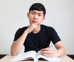 hombre asiático camisa negra cara seria y toca su barbilla con un libro en el escritorio mira la cámara en blanco aislado, concepto de tarea foto