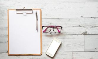 bolígrafo plateado en la hoja del documento portapapeles de madera con teléfono inteligente y gafas en la mesa de madera blanca vista superior espacio naturaleza sombra foto