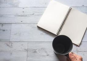 Atrapa la mano la taza de café de color negro con la página en blanco del cuaderno abierto en la mesa de madera blanca vista superior naturaleza sombra y concepto de idea de luz foto