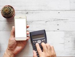 Hand holding cellphone white screen and using calculator with cactus and silver pen on white wood table nature shadow and sunlight top view and space business concept photo