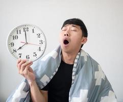 Young man wake up with blanket cover his body and feel sleepy yawn at face holding the clock in hand on white isolated photo