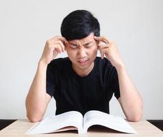 hombre asiático se esfuerza y toca su cabeza con dos manos con el libro sobre la mesa en blanco aislado, hombre serio con concepto de libro foto