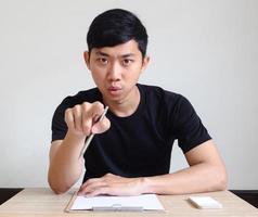 Asian man serious face point pen at you sit at the desk with board and mobile phone photo