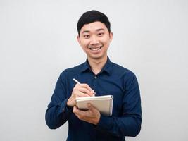 hombre asiático sonriendo y gesticulando escribiendo un cuaderno en la mano con fondo blanco foto
