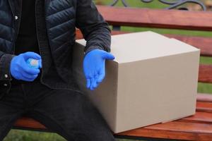 mensajero de servicio de entrega durante el coronavirus, covid-19, pandemia, mensajero recortado con máscara protectora médica, guantes rociando alcohol desinfectante en las manos cerca de cajas de cartón al aire libre foto