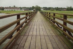 long wooden pedestrian bridge with railings across the river. background of village, srlective focus photo