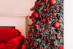 stylish christmas decorations on the tree, beautiful toys, balls in red colours. Big red bow on top. Xmas cozy atmosphere in living room, new year. Festive interior design. Happy holiday at home