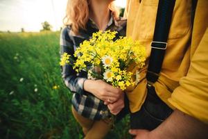 cropped photo of bouquet of wildflowers in woman's hand. Loving hipster couple walking in the field, kissing and holding hands, hugging, lying in the grass in the summer at sunset. valentines day