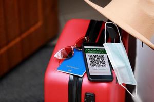 New normal concept. Close up view of a smartphone with an immune digital health passport, a red suitcase, protective mask, straw hat, passport and sunglasses. Traveling by plane during a pandemic. photo