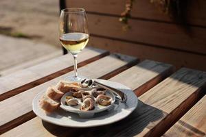 Glass with wine and dish with the snail shells, served on a white classic plate, with various sauces, pesto, truffle, cheese, pieces of French baguette, fork for a shell, wooden table in outdoor cafe photo