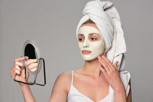 Beautiful female model with towel on her hear and white facial cosmetic mask on her face. Beauty cosmetic concept. Isolated on gray background. photo