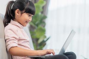 Asian Little school kid girl use laptop computer sitting on sofa alone at home. Child learning reading online social media content, play education lessons game chatting with friends. photo