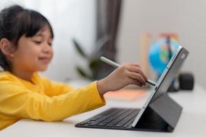 Asian schoolgirl doing her homework with digital tablet at home. Children use gadgets to study. Education and distance learning for kids. Homeschooling during quarantine. Stay at home photo