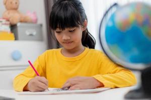 Asian schoolgirl doing her homework with digital tablet at home. Children use gadgets to study. Education and distance learning for kids. Homeschooling during quarantine. Stay at home photo