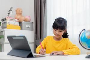 Asian schoolgirl doing her homework with digital tablet at home. Children use gadgets to study. Education and distance learning for kids. Homeschooling during quarantine. Stay at home photo