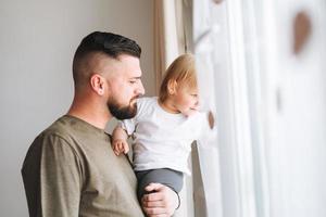 Multinational family, Young man father with baby girl on window sill looking at window at home photo