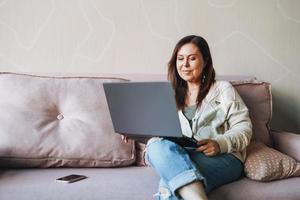 Adult brunette middle aged woman fifty years freelancer in casual clothes sing laptop sitting on couch at home photo