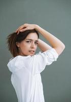 joven mujer infeliz con el pelo largo en camisa blanca sobre fondo gris, emoción negativa foto