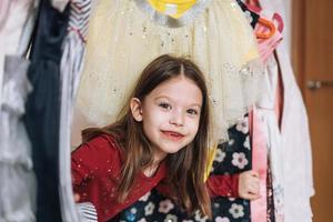 una niña sonriente con el pelo largo y oscuro vestido de rojo entre sus hermosos vestidos en el armario de la habitación de los niños en casa foto