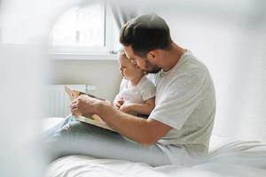 Happy father young man and baby girl little daughter having fun reading a book in children room at home photo