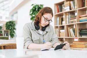 Young teenager girl in glasses doing homework with mobile phone at modern library public place, green open space office. University or college student listen online course in education class photo