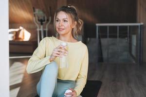 Young fitness blonde woman with bottle of water with lemon doing morning exercises near window of home photo