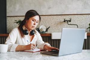 Adult smiling brunette woman in headphones doing notes in daily book with opened laptop at home photo