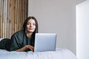 Beautiful smiling woman teenager girl student freelancer with dark long hair in casual using laptop sitting on bed at home photo