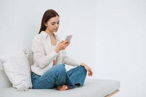Beautiful smiling woman teenager girl student with dark long hair in white shirt using mobile phone in hand sitting on couch at home photo