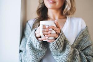 Close up portrait of Young beautiful woman forty year with blonde long curly hair in cozy knitted grey sweater with cup of tea in hands in bright interior at the home photo