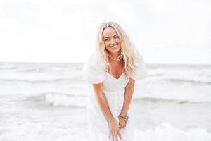 joven mujer rubia hermosa con el pelo largo en vestido blanco disfrutando de la vida en la playa del mar foto