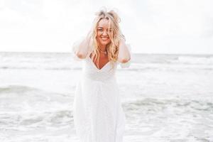 joven mujer rubia hermosa con el pelo largo en vestido blanco disfrutando de la vida en la playa del mar foto