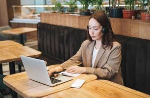 Adult thinking brunette woman forty years in headphones doing notes in daily book with opened laptop. She study something with online course at cafe photo